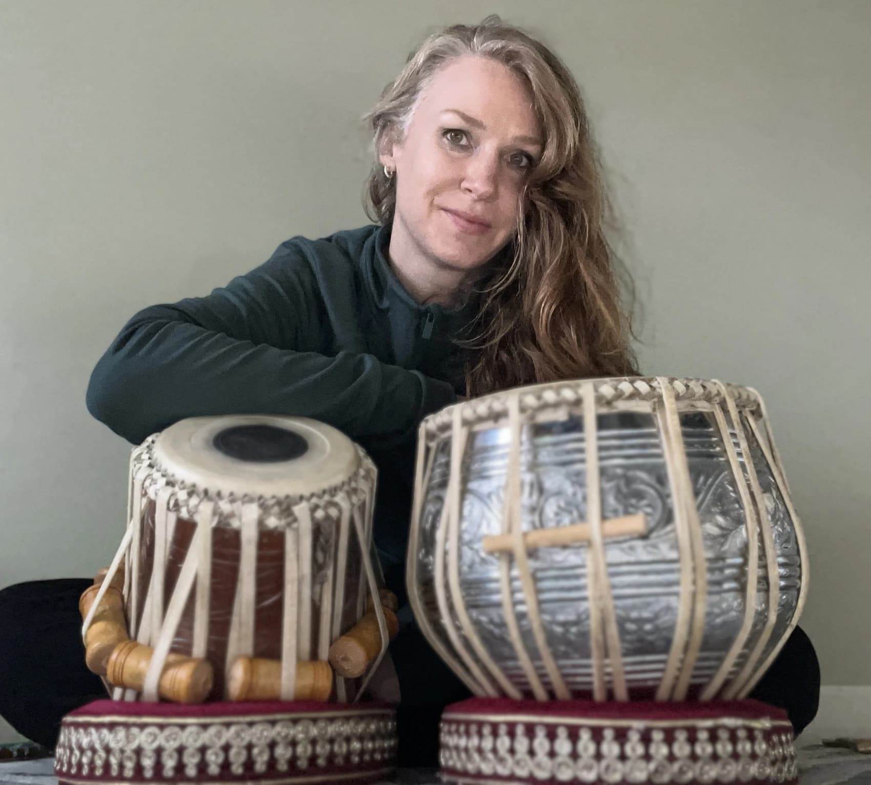 Melissa seated behind her tabla against a plain wall in soft light.